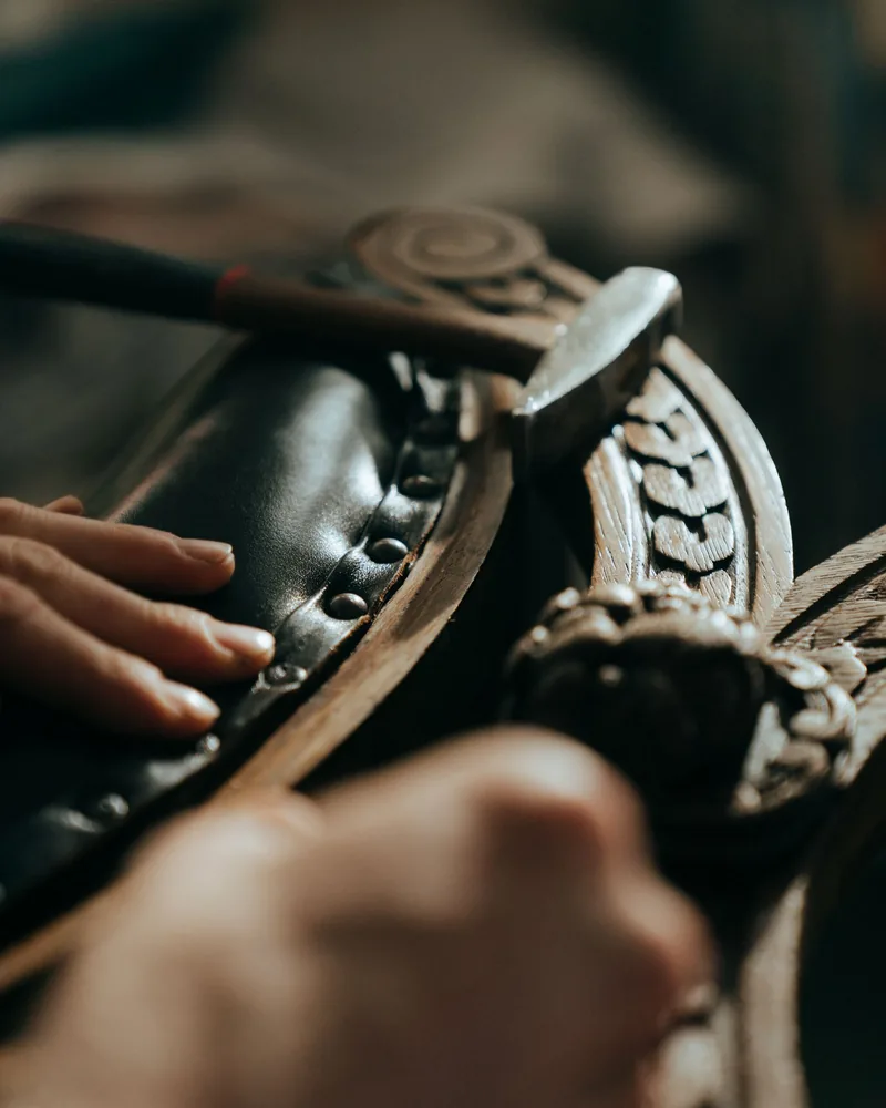 Detalle de una talla de madera y una tapicería con clavos decorativos durante una reparación, con un martillo de tapicero apoyado sobre la pieza